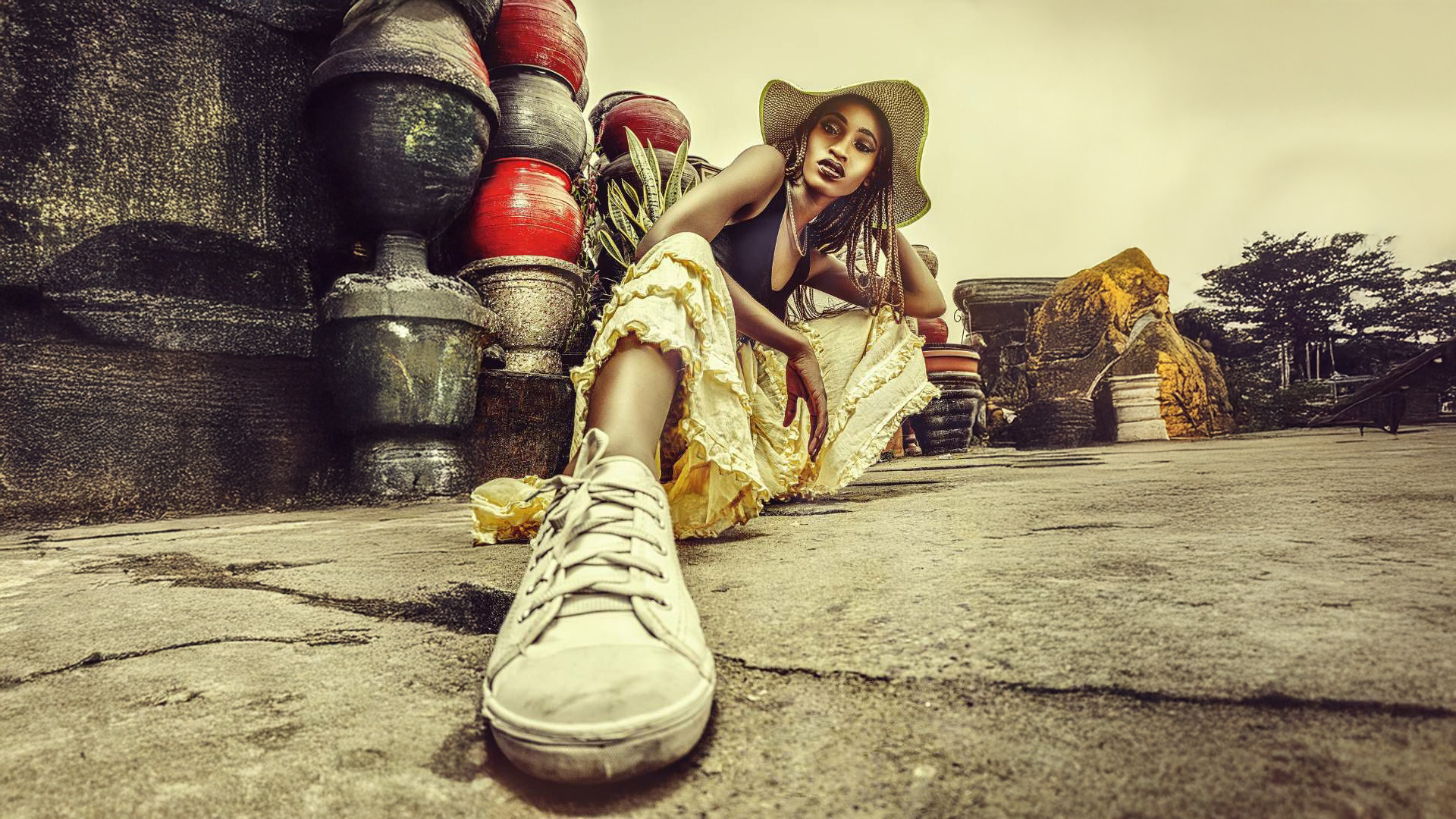 Fashion photograph of a model in a flowing yellow ruffled dress and wide-brimmed hat, seated on the ground with one leg extended to emphasize a white sneaker. The industrial backdrop features stacked red and gray containers, creating a striking contrast with the feminine styling. The image uses dramatic lighting and a vintage-inspired color palette.