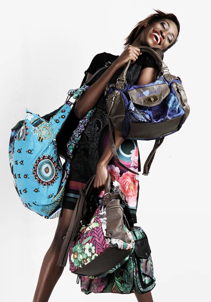 Professional fashion photograph showing a model in a black dress against a white background, playfully posed while holding multiple designer bags. She wears a short black dress and displays four distinctive handbags: a large turquoise bag with circular patterns, a navy and grey structured bag, and two floral-patterned bags in pink and green tones. The model's expression is cheerful and energetic, with her head tilted back in laughter.